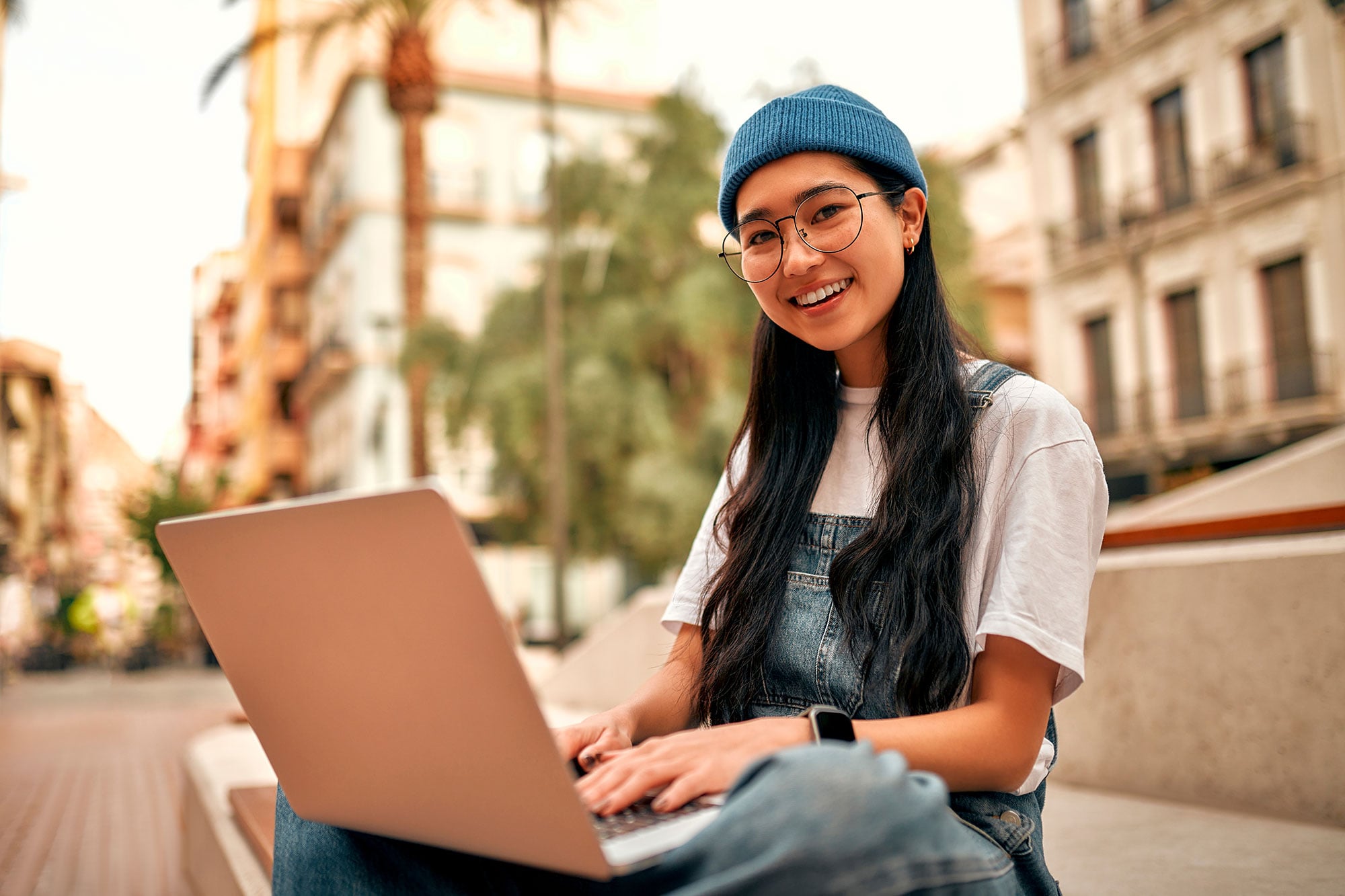 Smiling adult in beanie and overalls typing on a laptop outdoors.